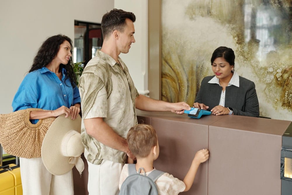 Une famille (parents et enfant) arrive à une réception, l'homme utilisant une carte pour payer ou s'enregistrer auprès de la concierge, illustrant un accueil de voyageurs.