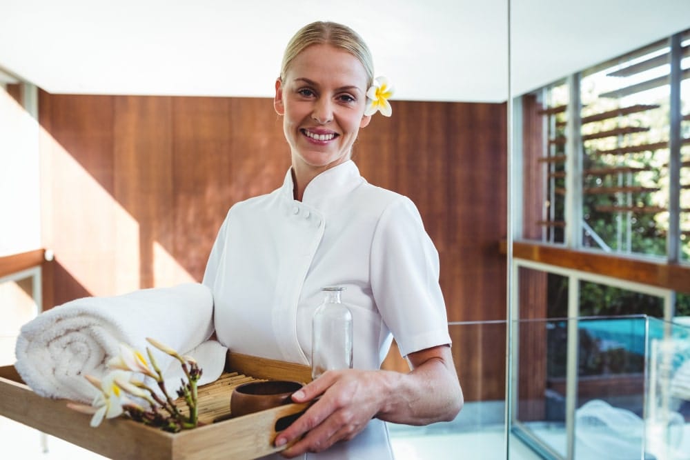Femme blonde en uniforme de spa (massage/esthétique) souriant et tenant un plateau de service avec serviettes blanches, huile et fleur de frangipanier, dans un intérieur moderne et boisé.