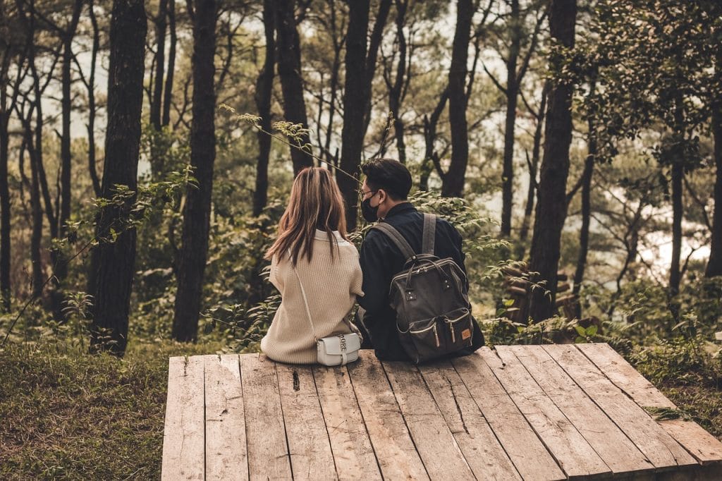 Vue de dos d'un couple (homme et femme) assis côte à côte sur une plateforme en bois, observant le paysage d'une forêt de pins dense et ombragée.