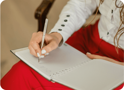 Une main de femme manucurée écrivant avec un stylo argenté dans un carnet à spirale, vêtue d'un chemisier blanc et d'un pantalon rouge.