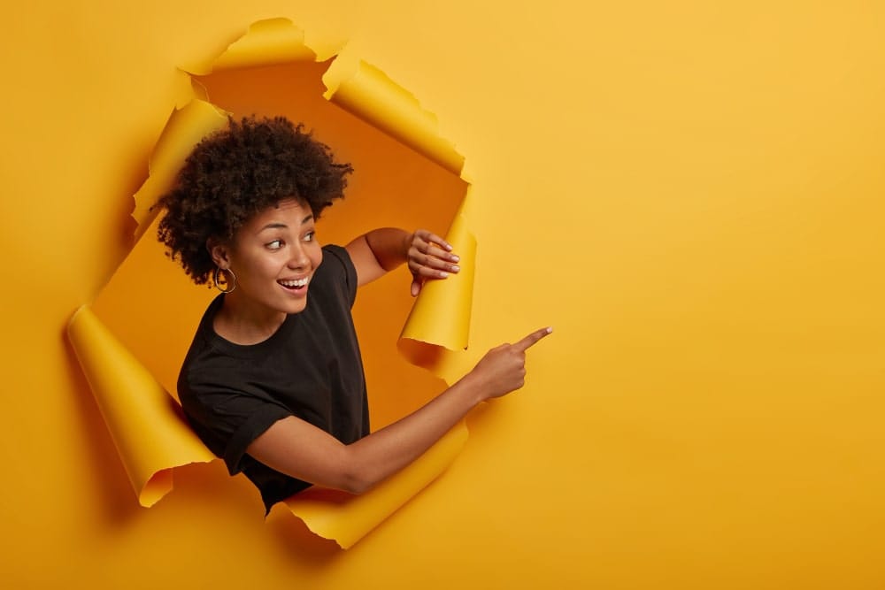 Une femme noire, souriante et surprise, sort d'un trou déchiré dans un mur en papier jaune, pointant l'espace vide à droite pour y insérer un texte promotionnel.
