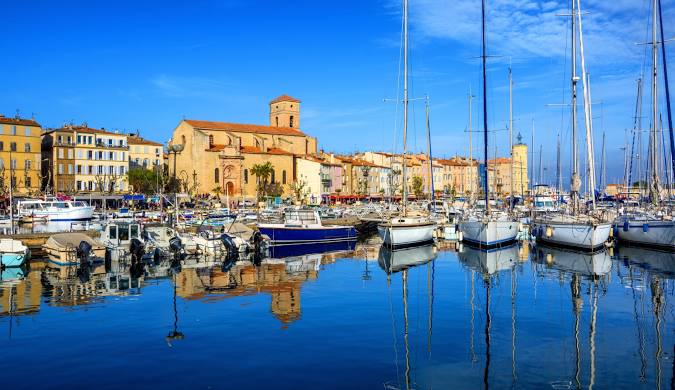 Vue panoramique du vieux port de Saint-Tropez ensoleillé, avec des voiliers amarrés, le clocher ocre de l'église et les façades colorées reflétées dans l'eau bleue.