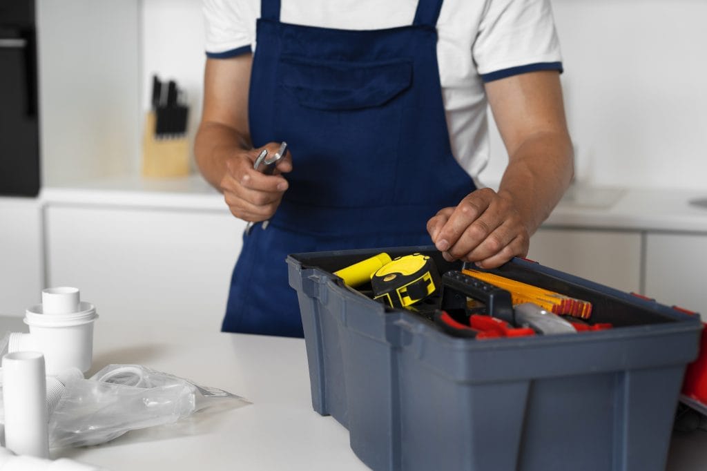 Plombier en salopette bleue préparant ses outils dans une cuisine blanche, avec une boîte à outils remplie et des raccords de plomberie à côté.