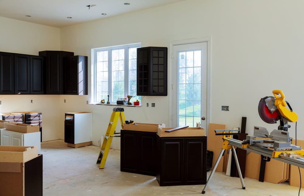Cuisine en cours de rénovation avec armoires foncées installées, fenêtre, porte et scie à onglet professionnelle.
