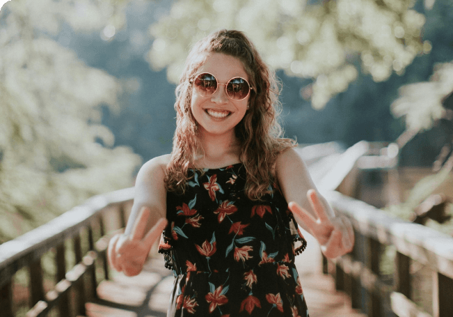 Jeune femme souriante aux cheveux bouclés portant des lunettes de soleil rondes et un chemisier fleuri, faisant le signe V de la victoire sur une passerelle en bois en forêt.