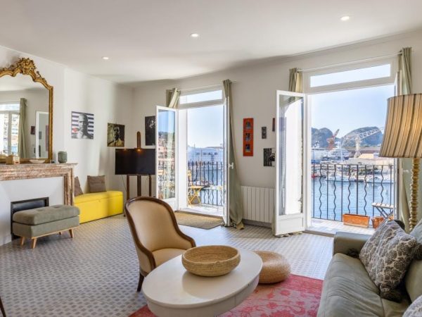 Salon lumineux de style classique avec cheminée, miroir doré, et vue panoramique sur un port industriel depuis le balcon.
