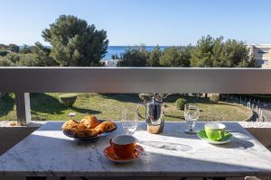 Petit-déjeuner sur une table en marbre sur un balcon, avec croissants et vue sur la mer Méditerranée et les pins.